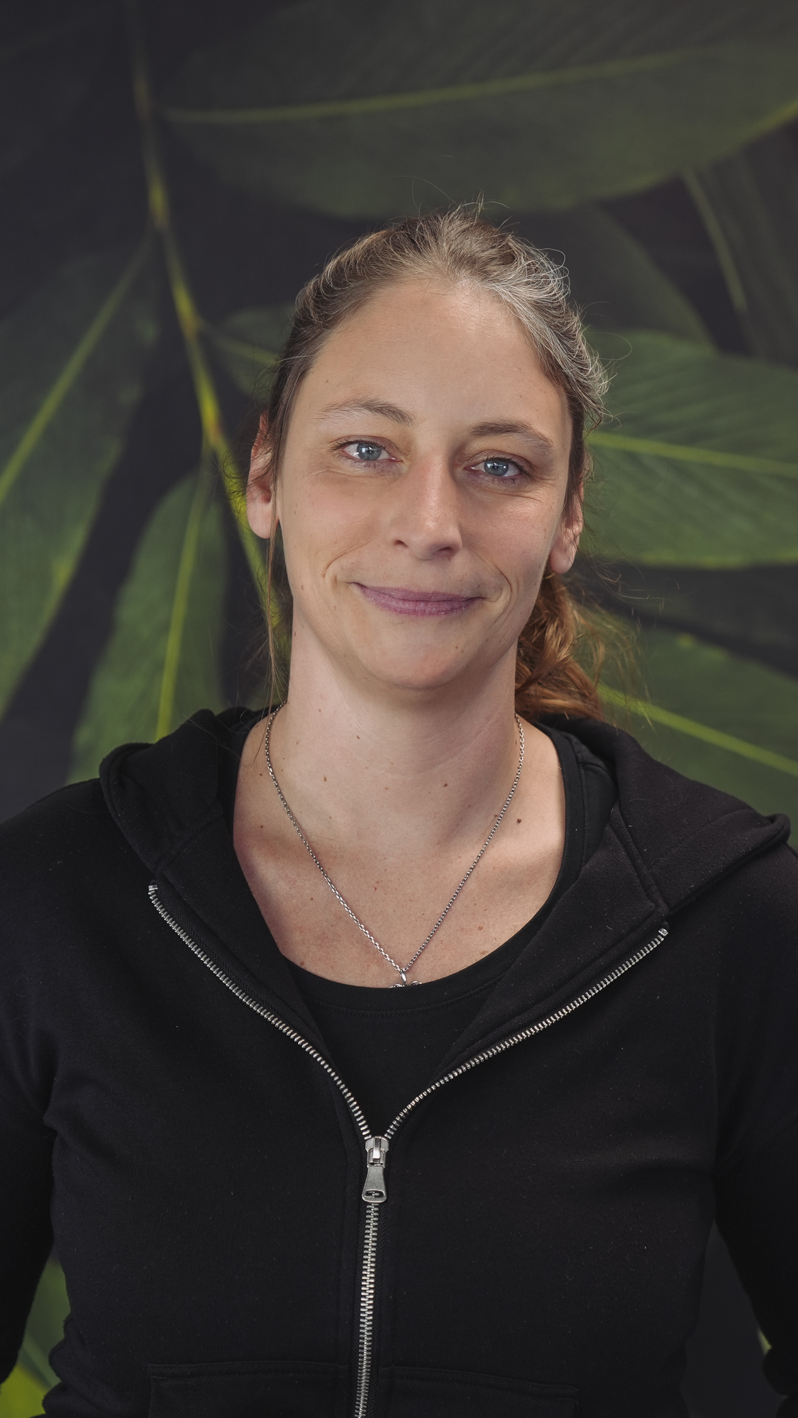 A person in a black hoodie standing against a leafy background, smiling slightly.