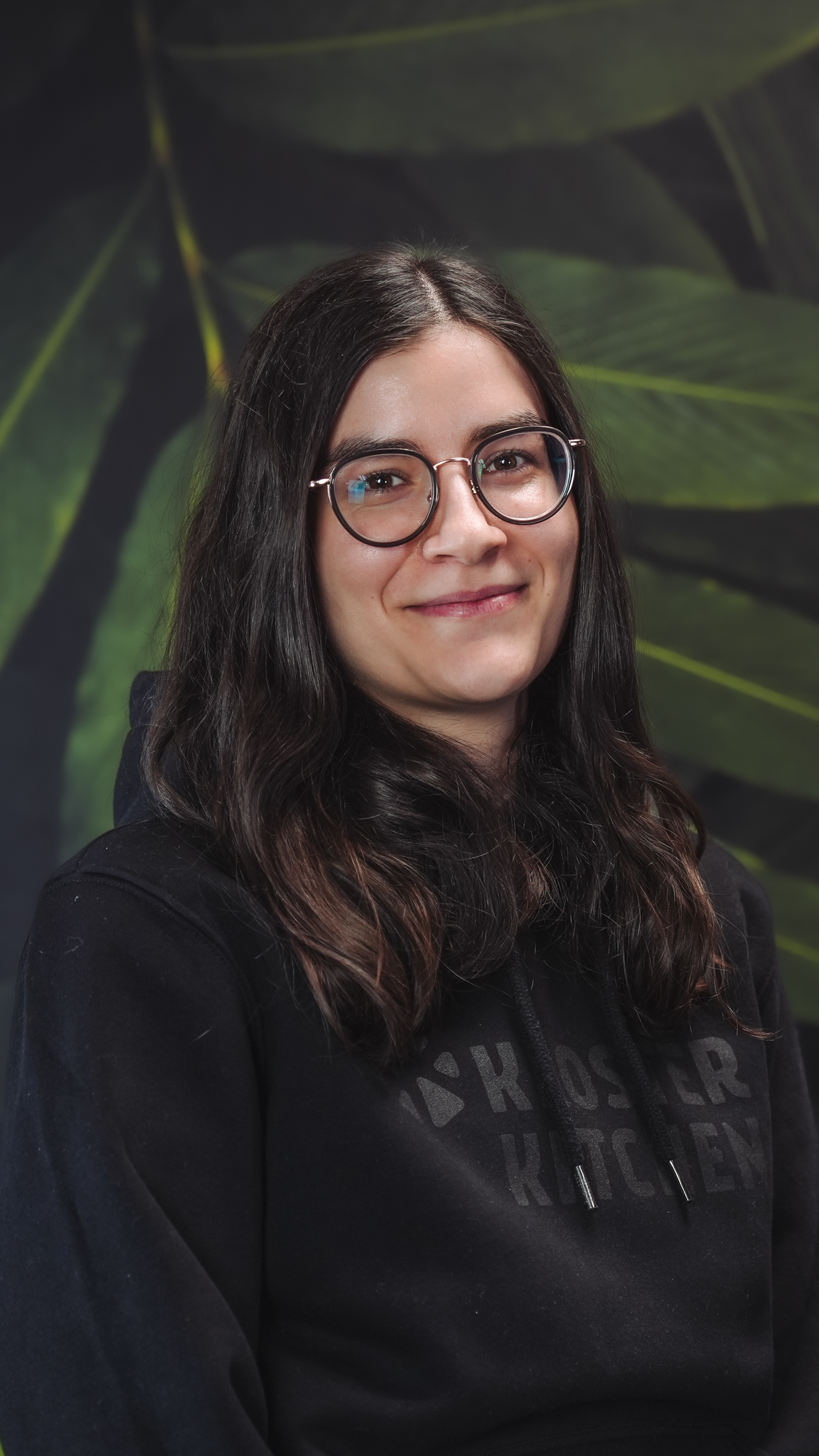 Person smiling in front of a leafy background, wearing glasses and a black hoodie.