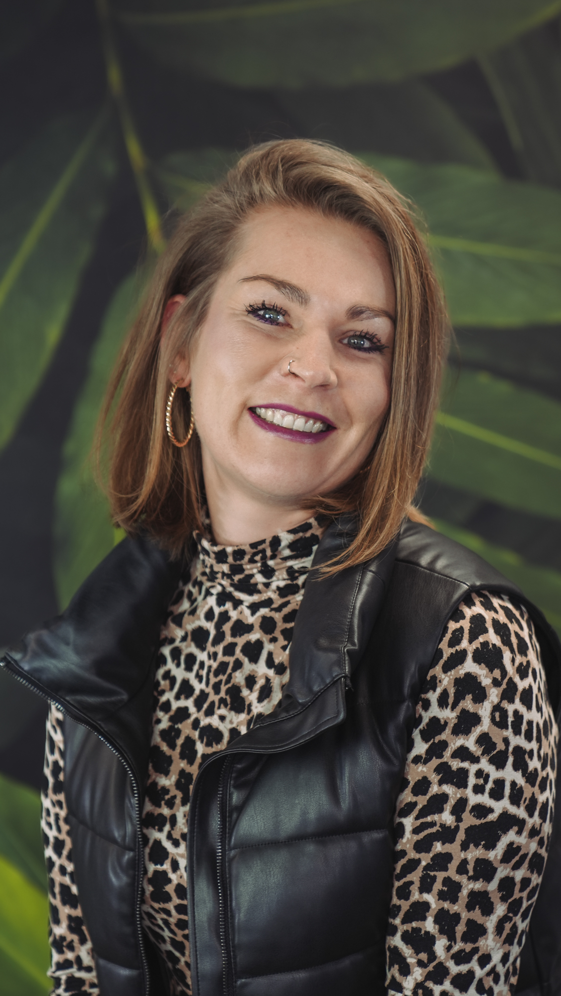 Smiling person wearing leopard print top and black vest, with a leafy background.
