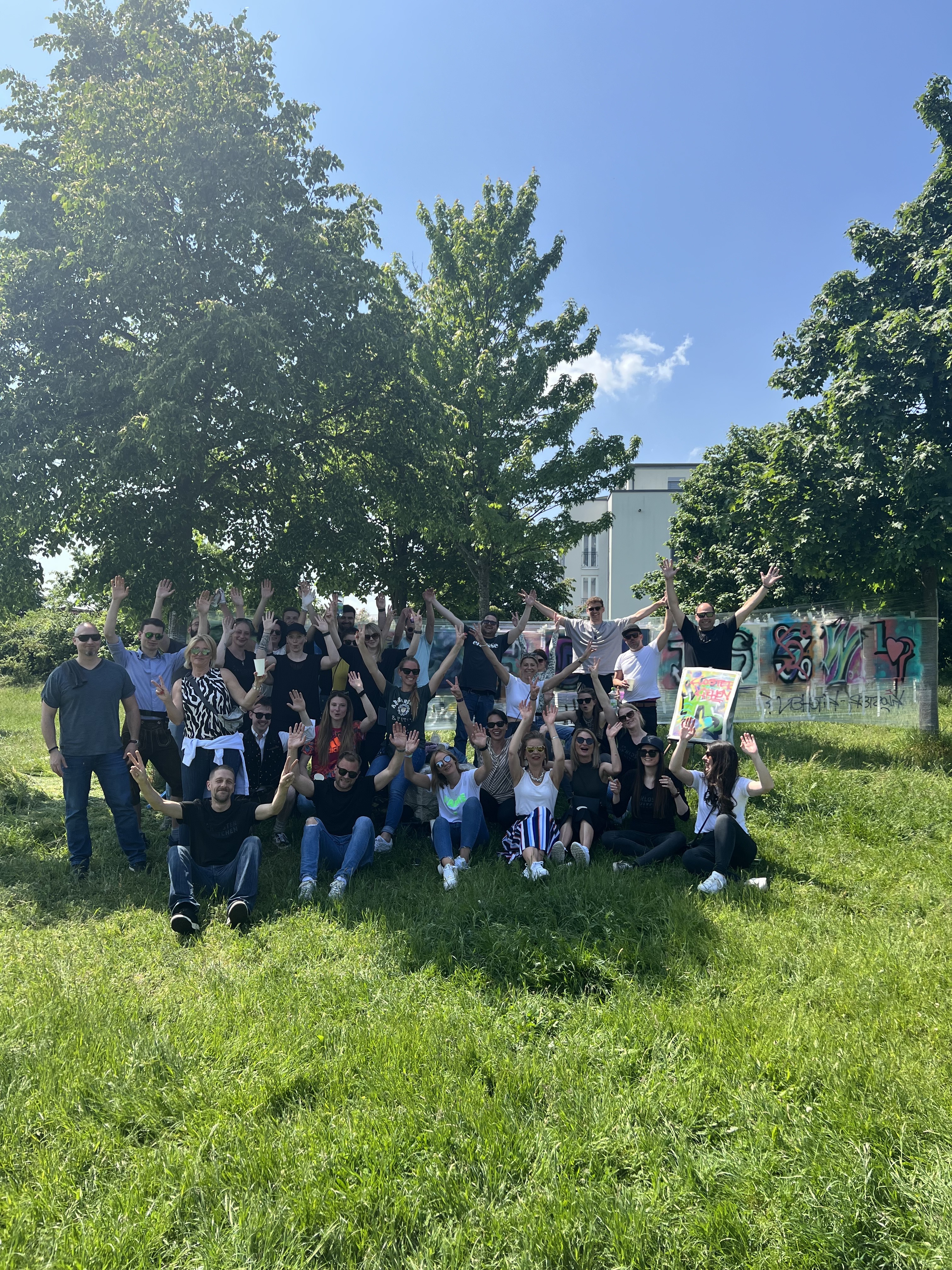 A large group of people gathers outdoors, smiling and waving in a park setting.