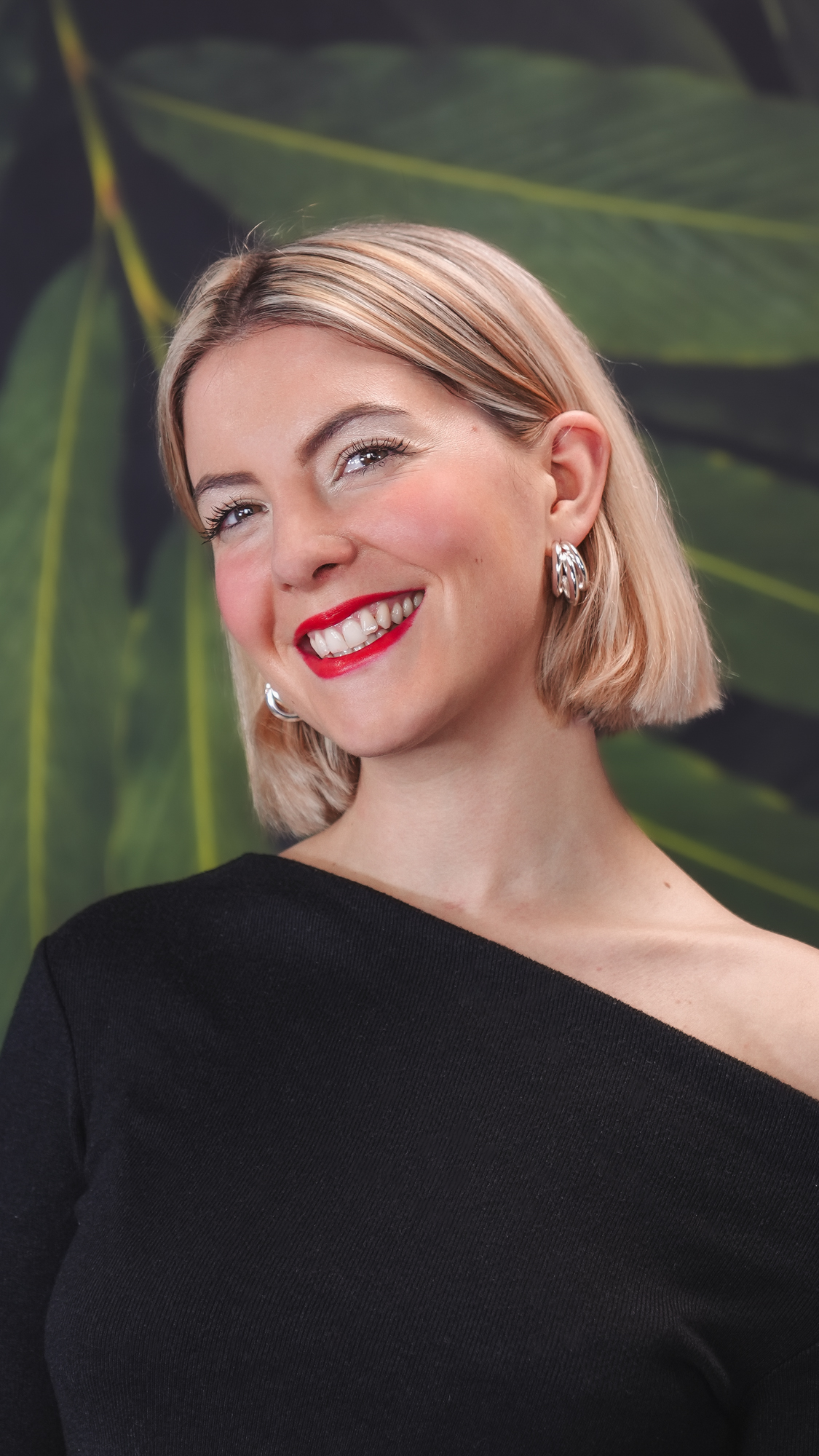 A woman in black top smiling in front of green leaves background.
