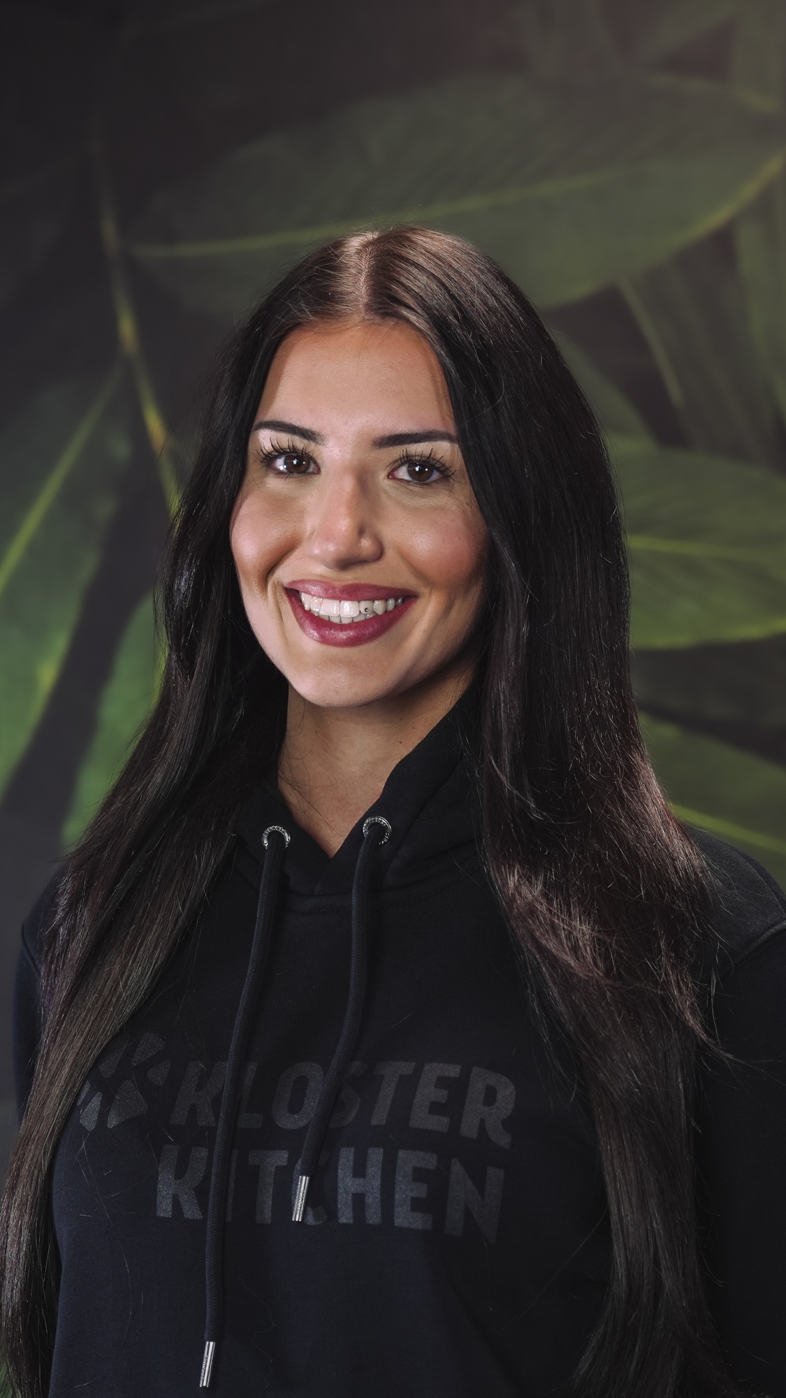 Smiling person with long dark hair, wearing aKloster Kitchen' hoodie with a leafy background.