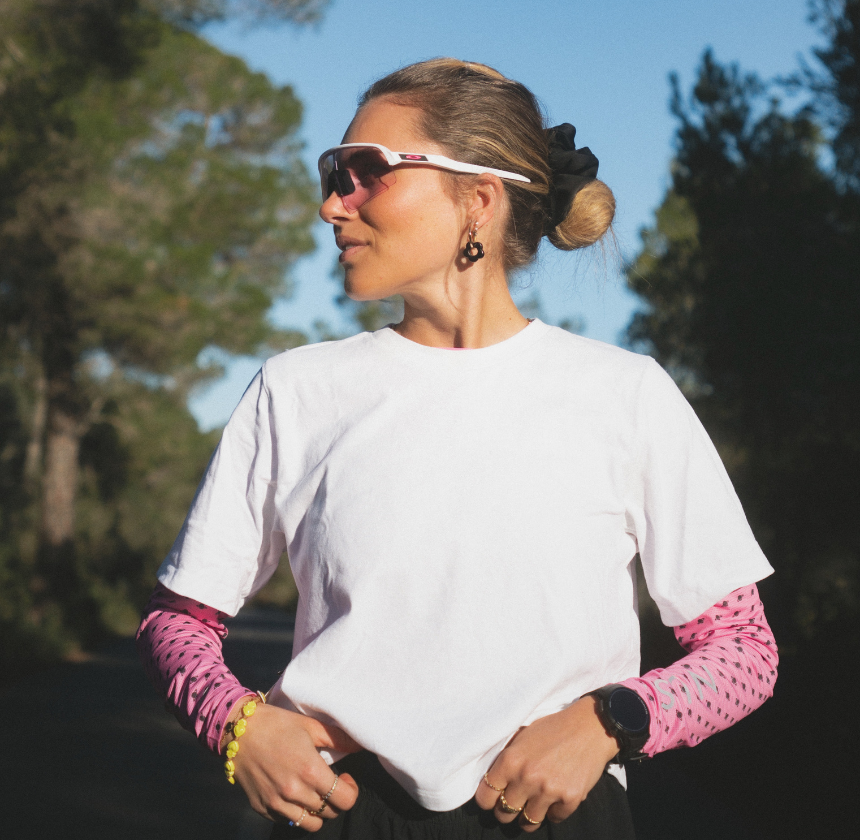 Person in sunglasses and white shirt outdoors, wearing pink sleeves and a watch.