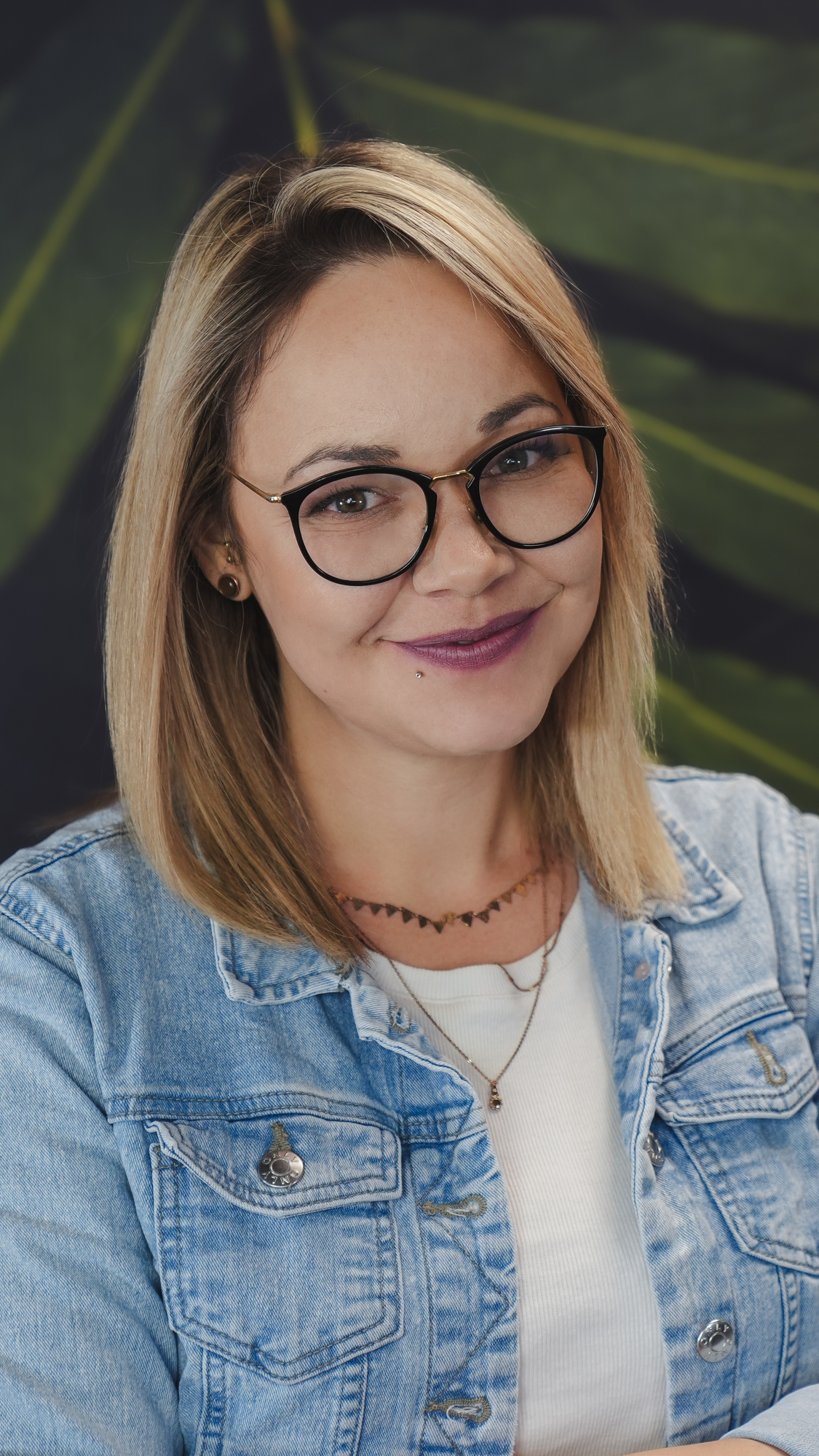 Woman smiling in glasses and denim jacket.