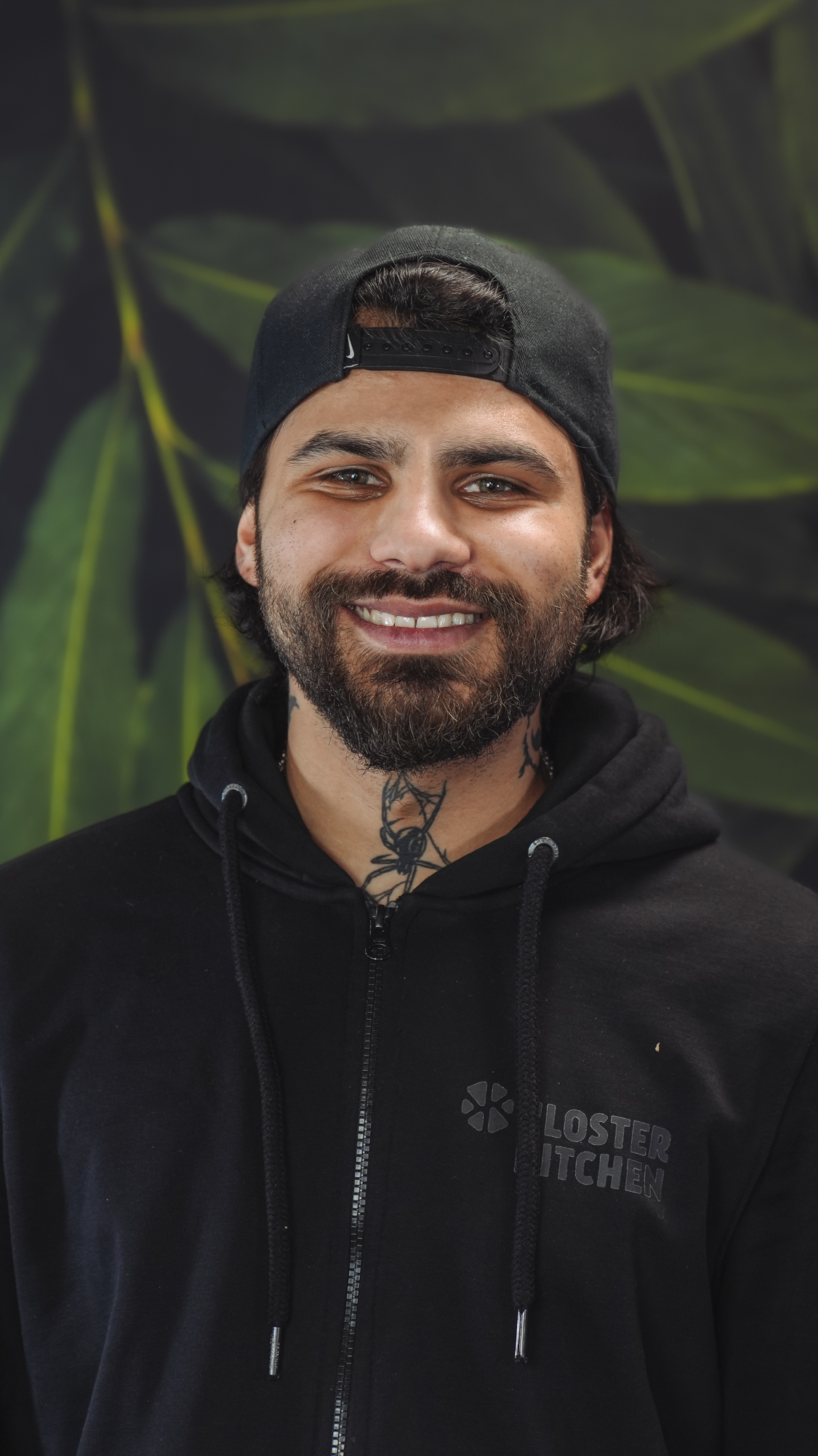 Man with a beard and cap smiling, standing against a leafy background.