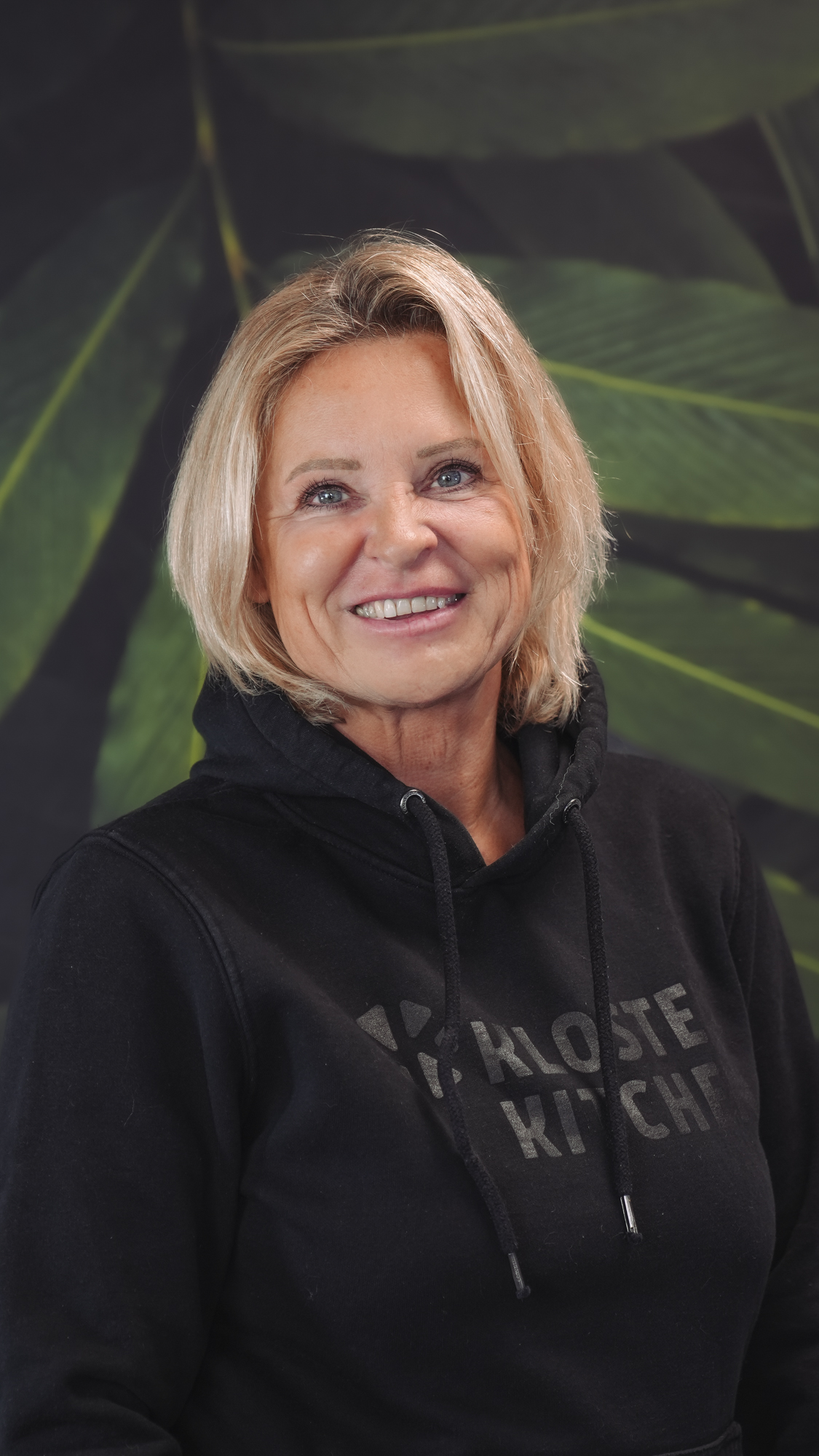 Woman in black hoodie smiling, standing against a leafy green background.