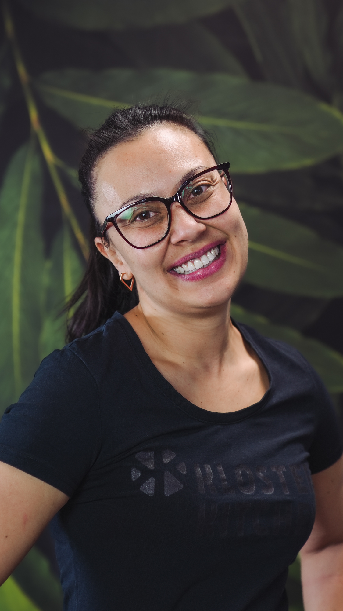 Smiling woman with glasses and black shirt, in front of leafy background.