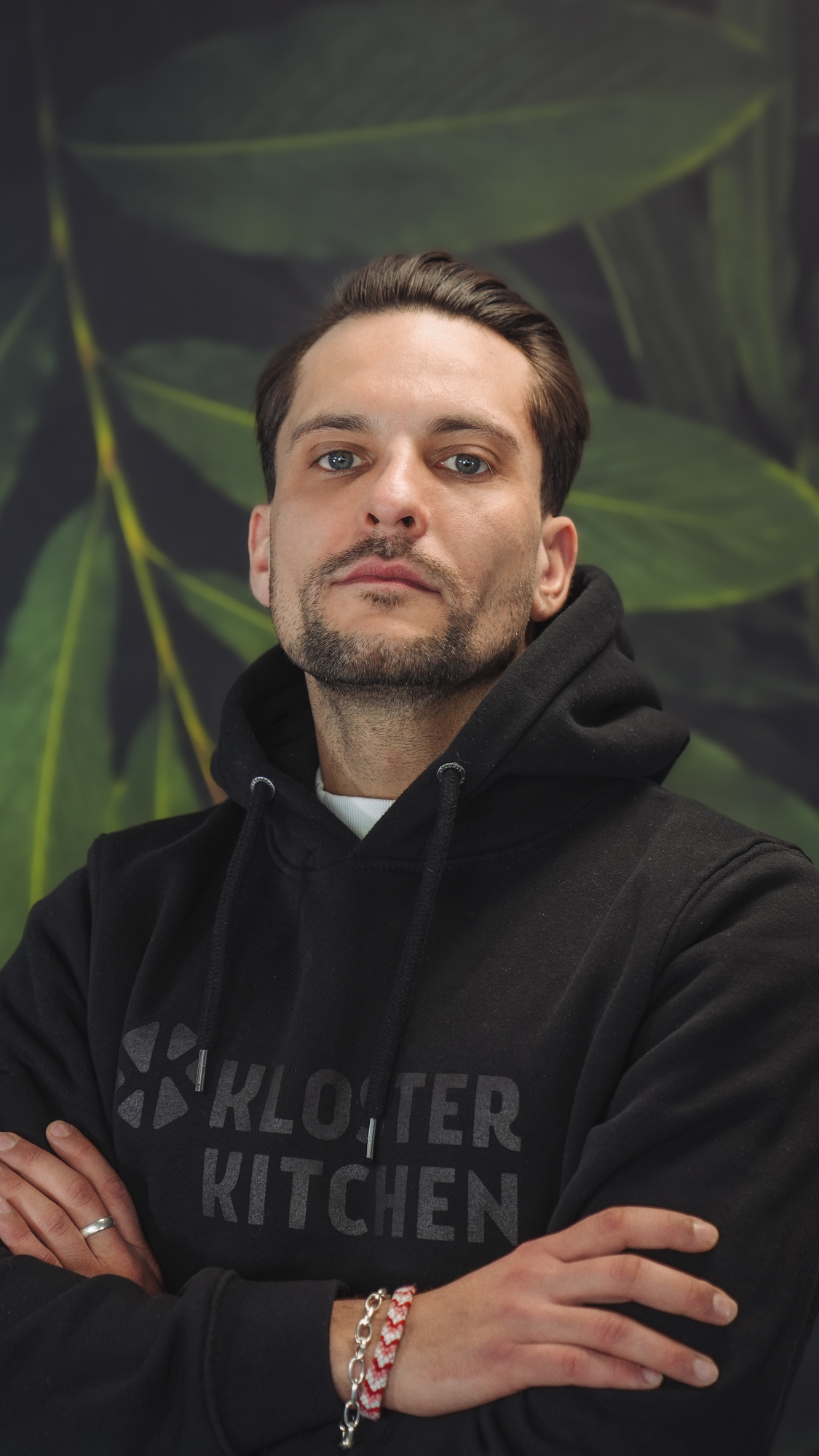 A person in a black hoodie with arms crossed against a leafy background.