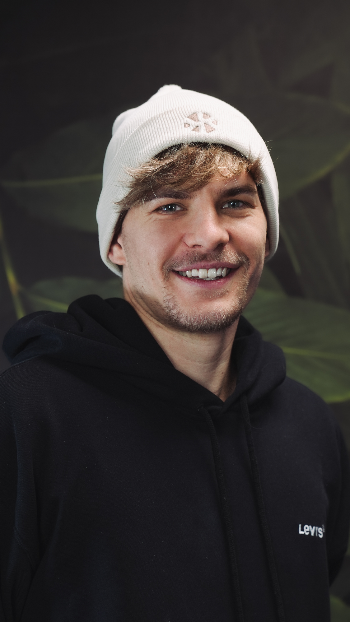 A person wearing a white beanie and black hoodie, smiling against a leafy background.