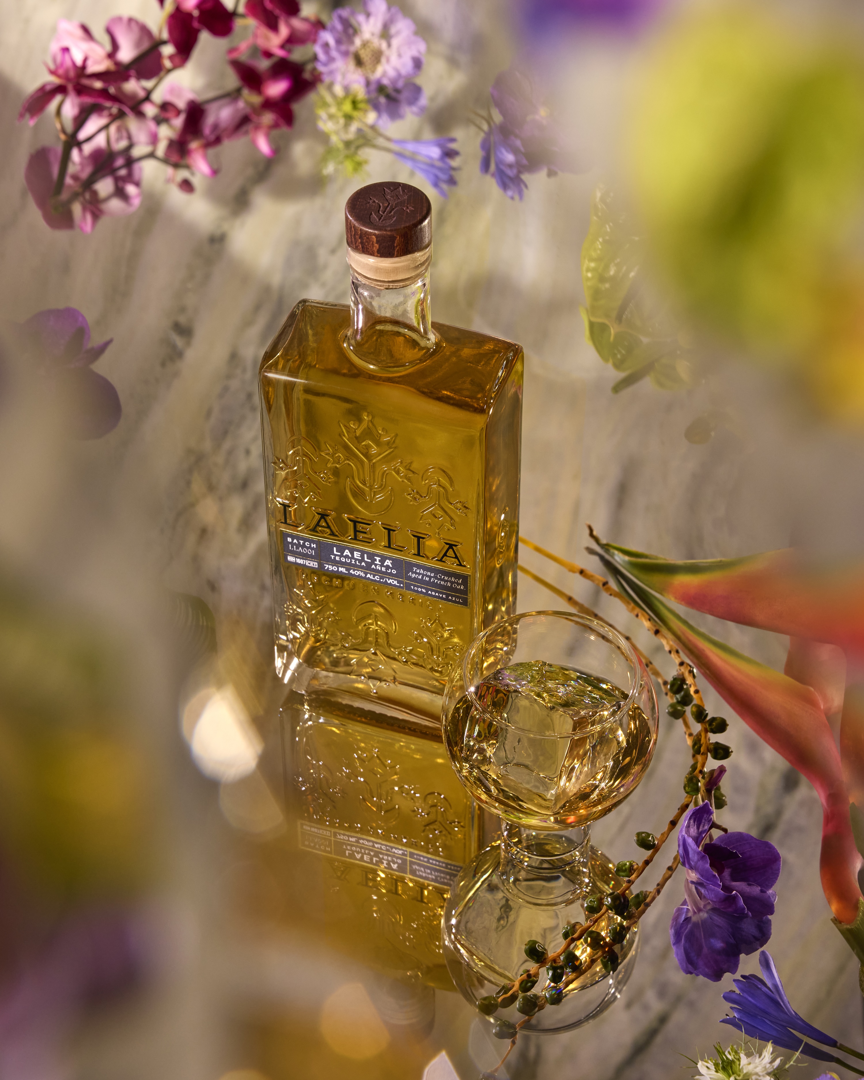 Bottle of amber liquid with flowers and a glass on a reflective surface.