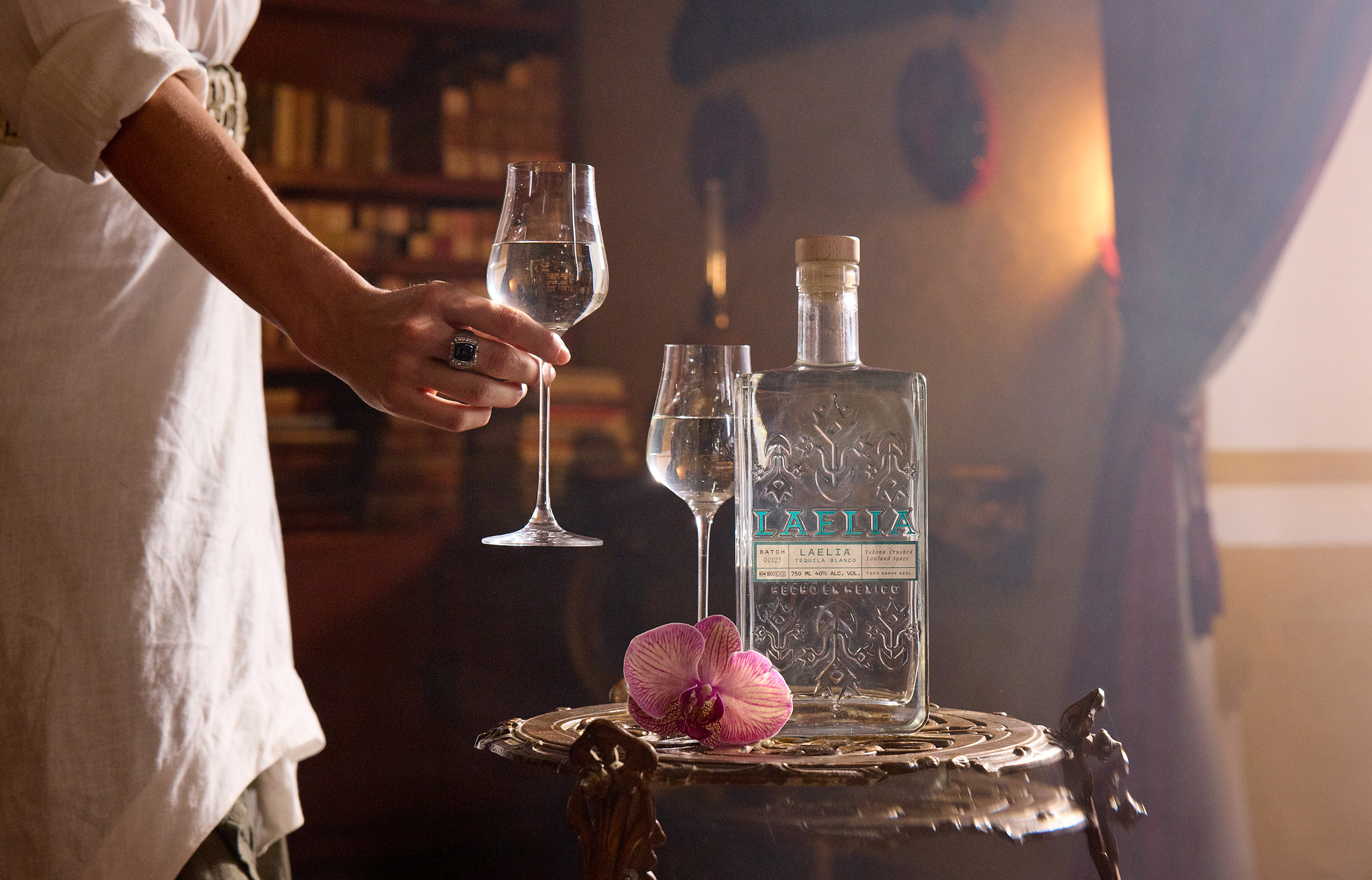 Person holding a wine glass near a bottle on a table with a flower.