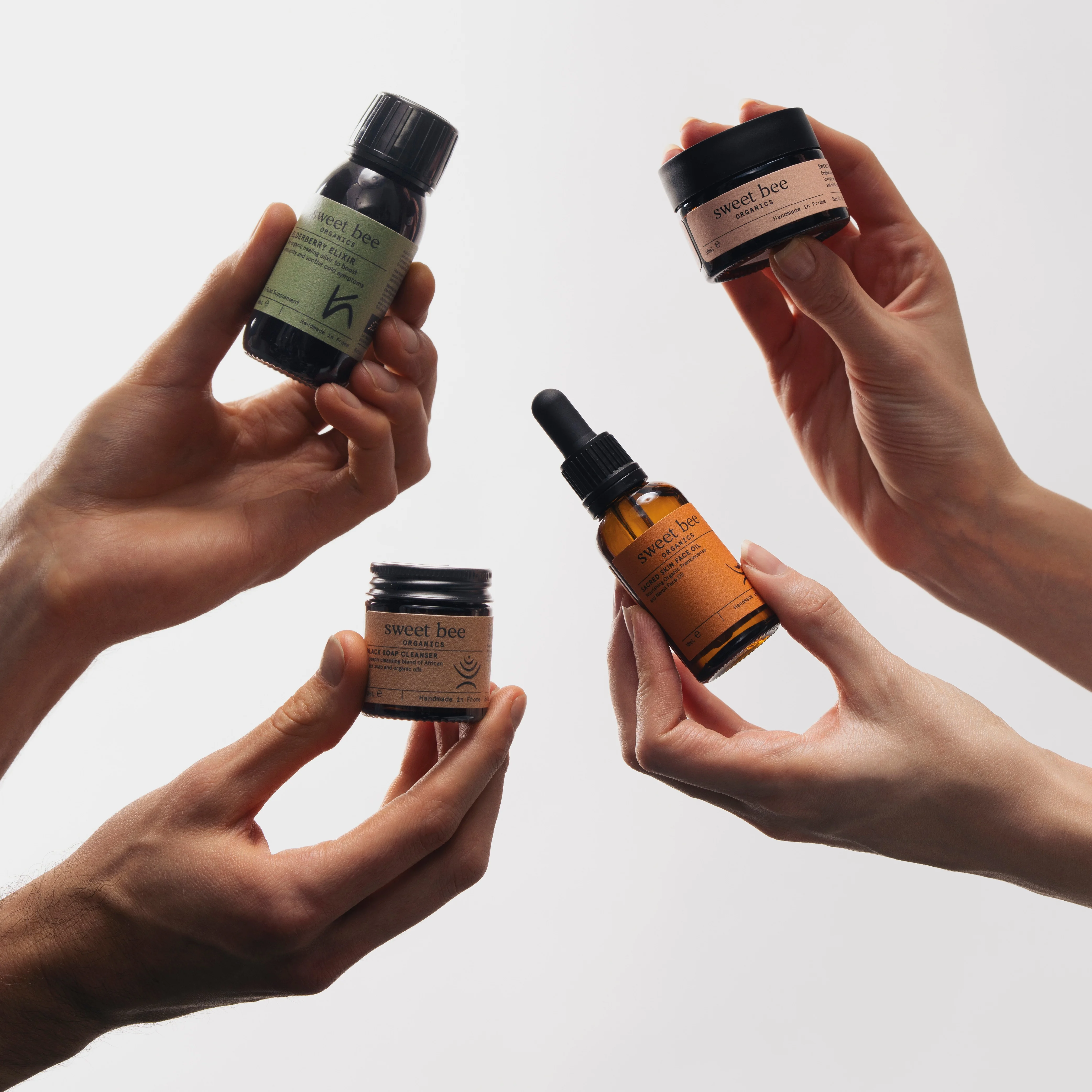 Four hands holding up various small jars and bottles of Sweet Bee Organics products against a white background.