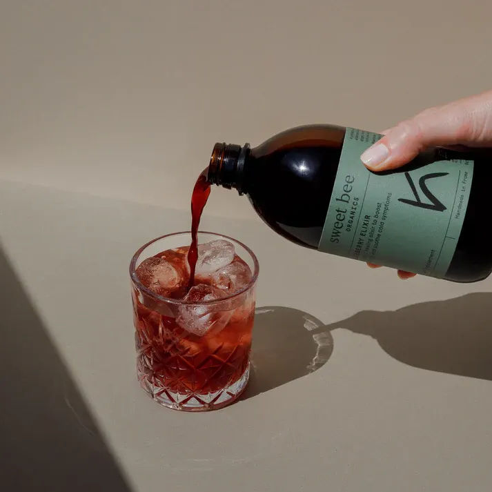 A hand pours a red beverage from a dark bottle into a decorative glass filled with ice.