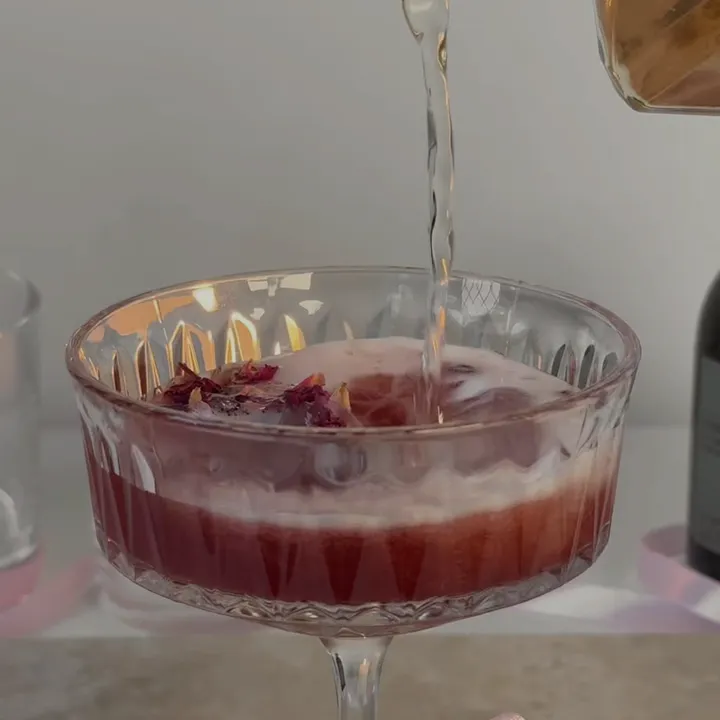A clear liquid is being poured into a pink cocktail in a coupe glass, garnished with rose petals.