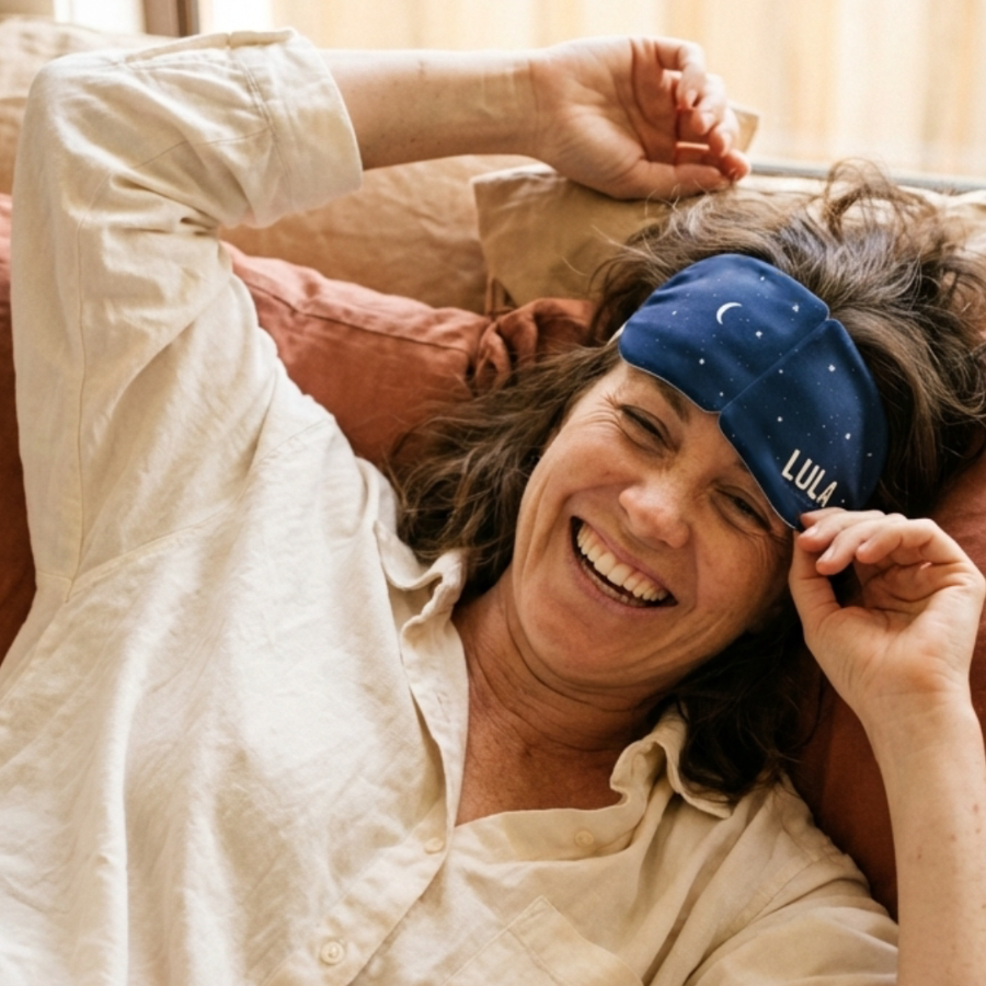 A smiling woman in bed lifts a personalized blue sleep mask with the name Lula on it.