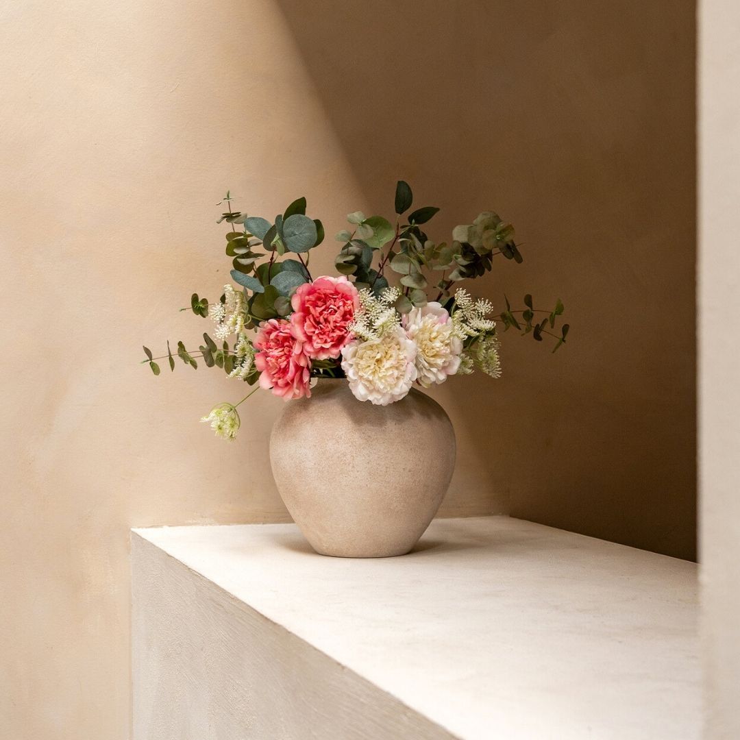 A textured beige vase with pink and cream flowers and greenery sits on a white ledge.
