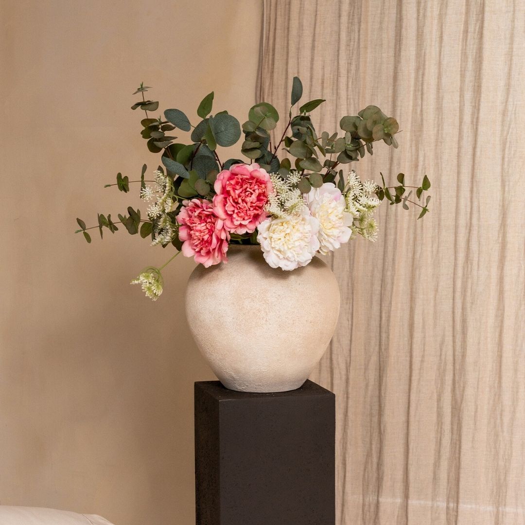 A bouquet of pink and white flowers with greenery in a textured beige vase on a black pedestal.