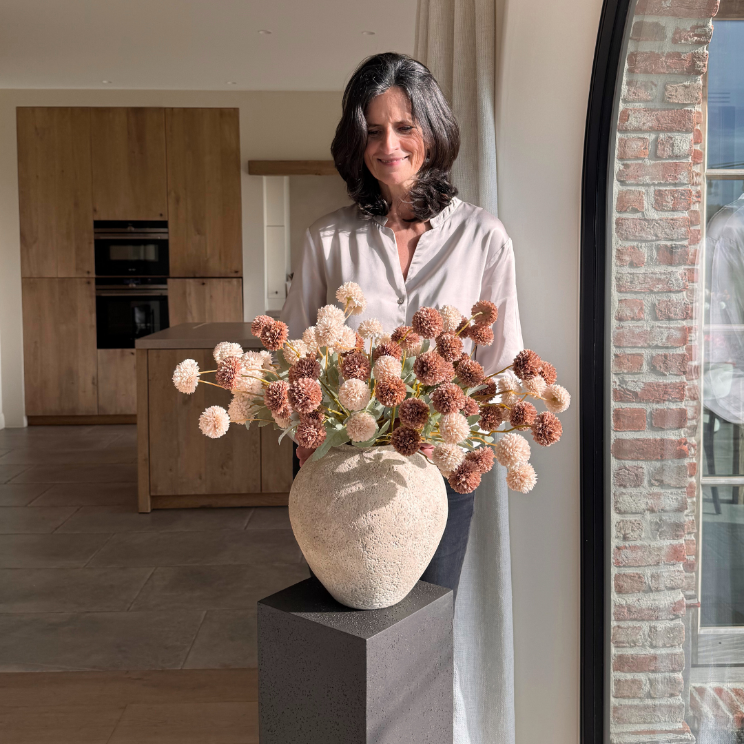 A woman stands in a sunny room, holding a large vase filled with pink and brown pom-pom flowers.