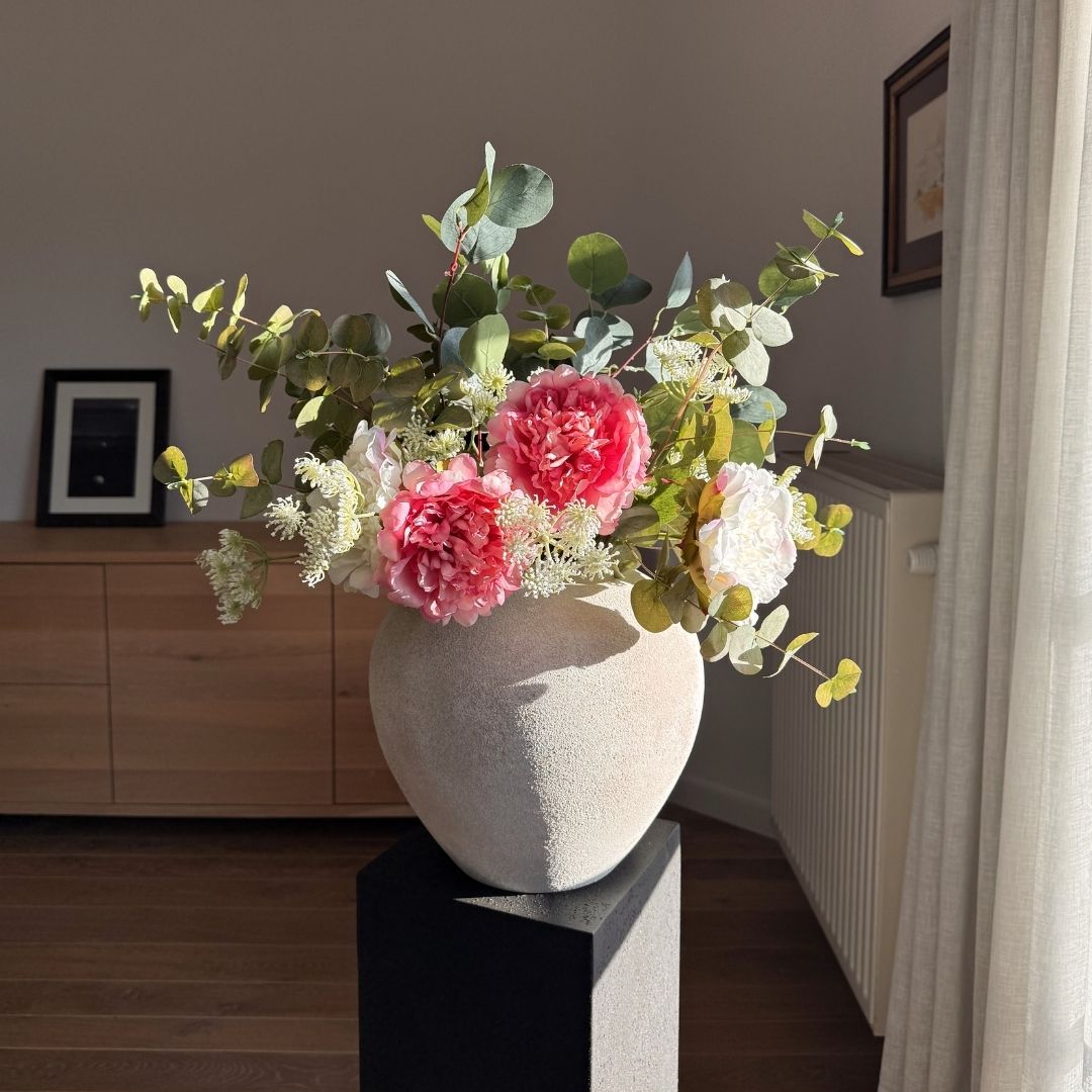 A textured vase with pink and white flowers and eucalyptus leaves sits on a black pedestal in sunlight.