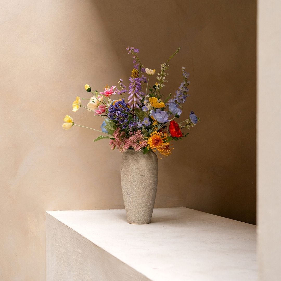 A tall, textured vase holding a colorful arrangement of wildflowers sits on a white ledge against a tan wall.