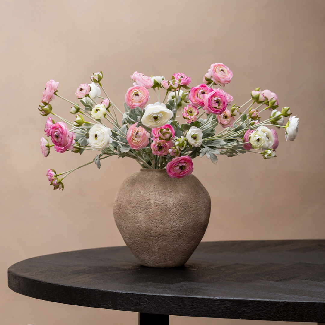 A bouquet of pink and white ranunculus in a textured brown vase on a dark wood table.