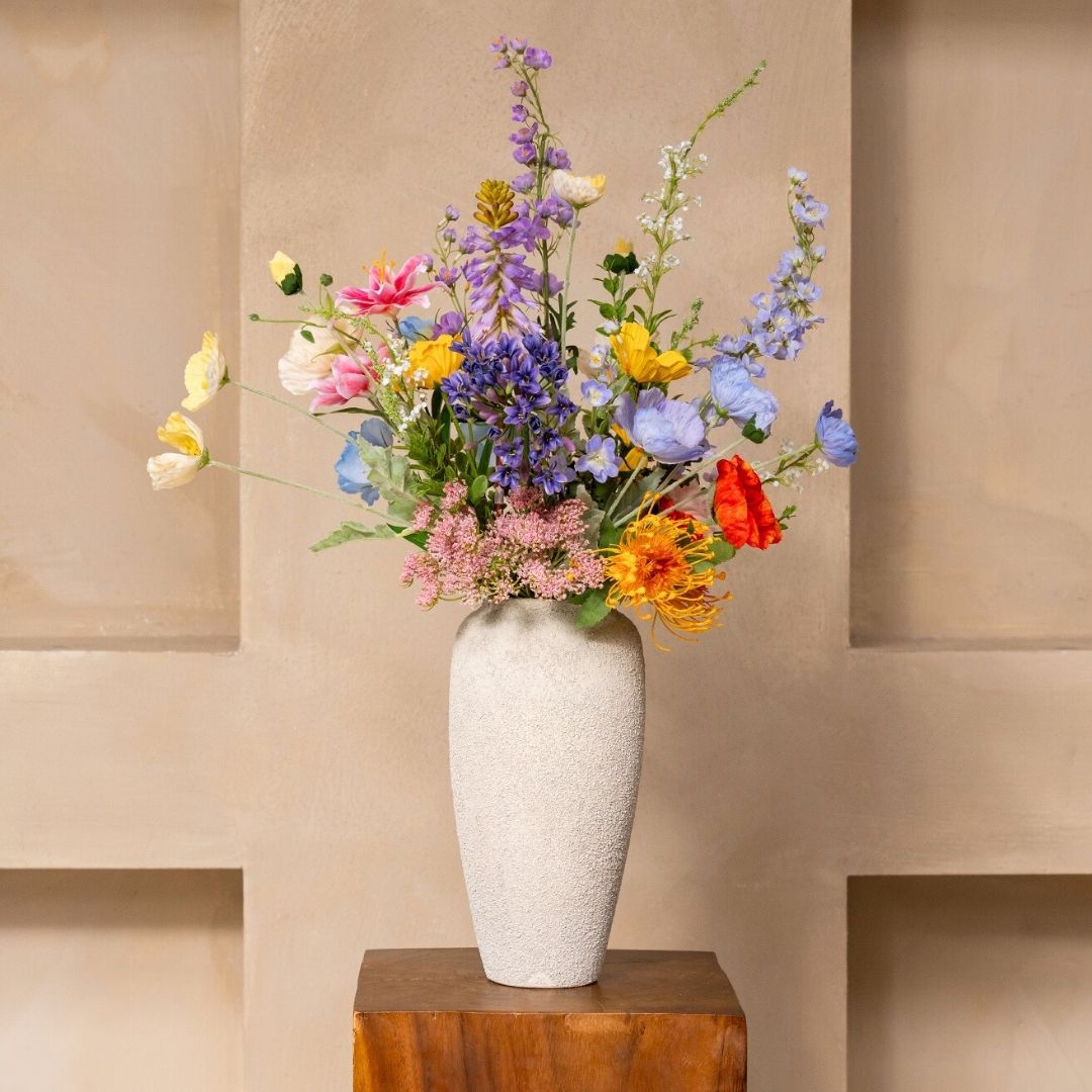 A tall, textured white vase filled with a colorful arrangement of wildflowers sits on a wooden pedestal.