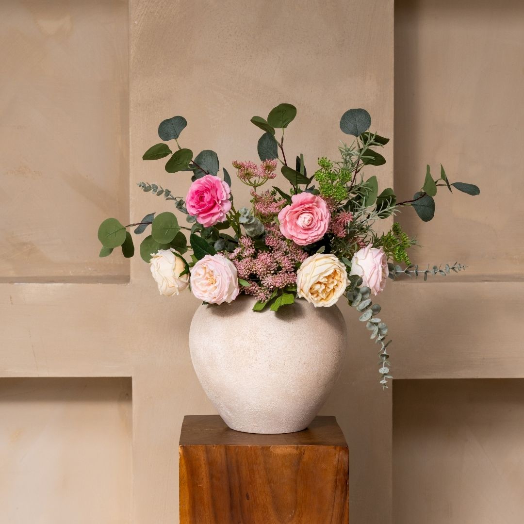 A bouquet of pink and white roses with greenery in a round, textured vase on a wooden stand.