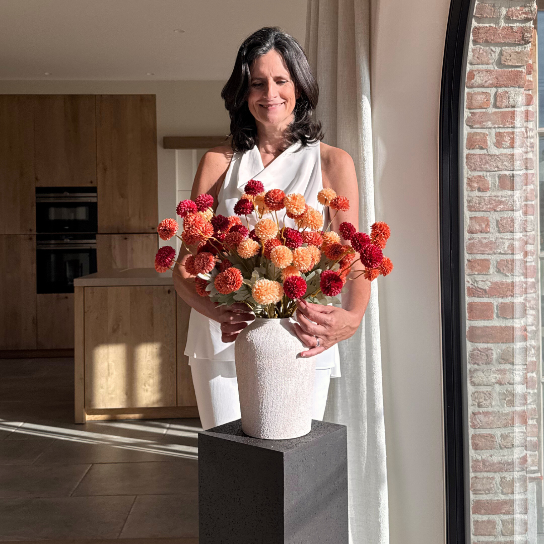 A woman in a white outfit arranges a bouquet of red and orange pom-pom flowers in a vase.