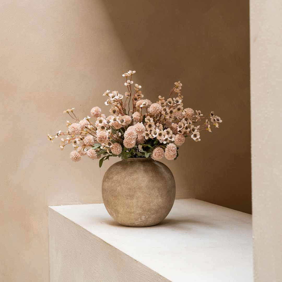 A round, rustic vase holding a bouquet of small, light pink flowers on a white ledge.