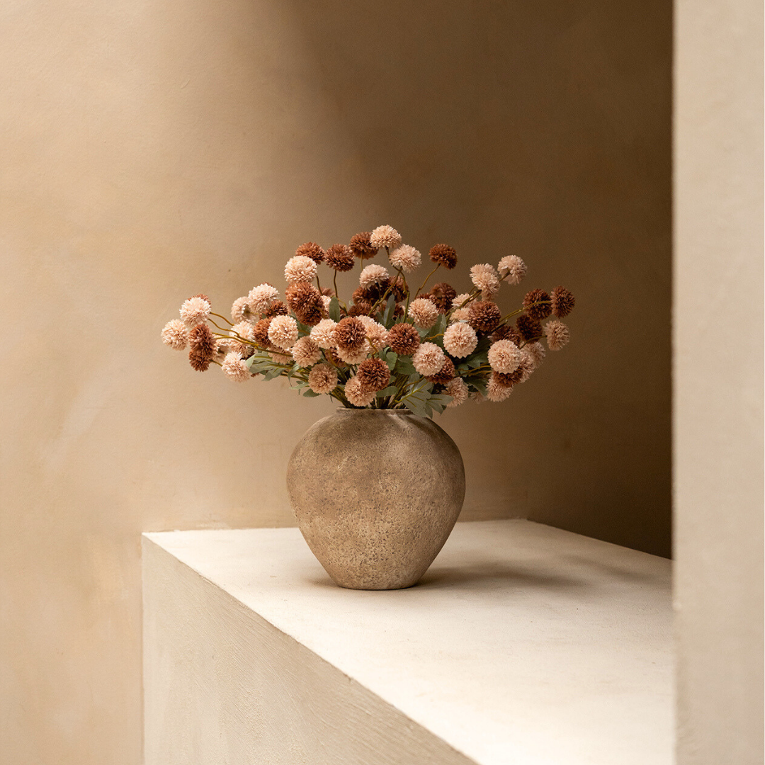 A stone-textured vase filled with light and dark brown pom-pom flowers sits on a ledge.
