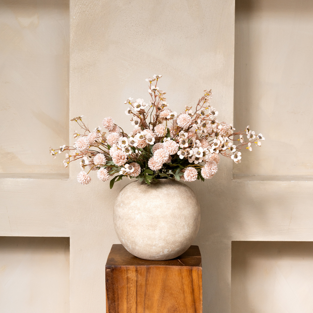 A bouquet of pink and white flowers in a round, textured vase sitting on a wooden pedestal.