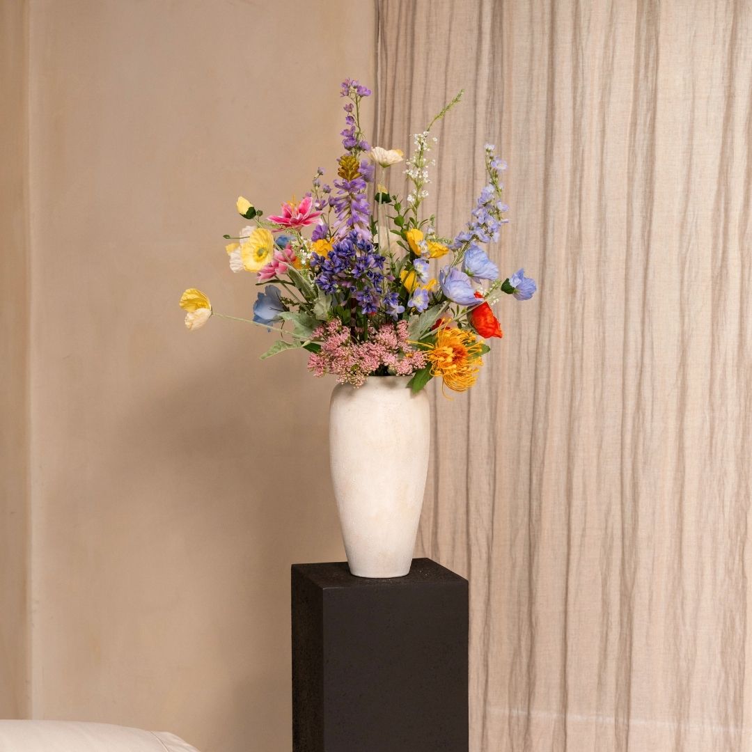 A tall white vase filled with colorful wildflowers sits on a black pedestal in front of a curtain.