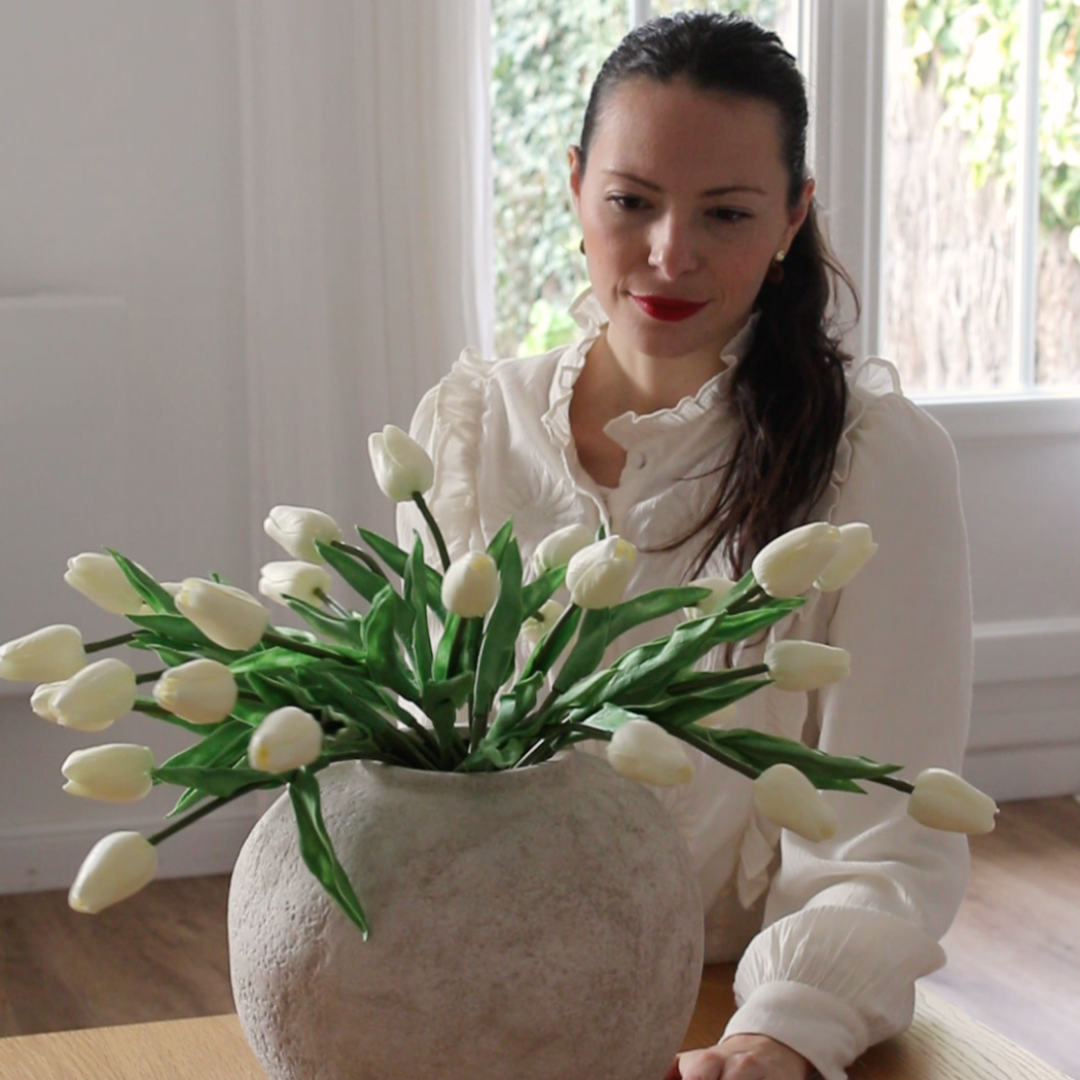 A woman in a white blouse looks down at a large vase filled with white tulips.