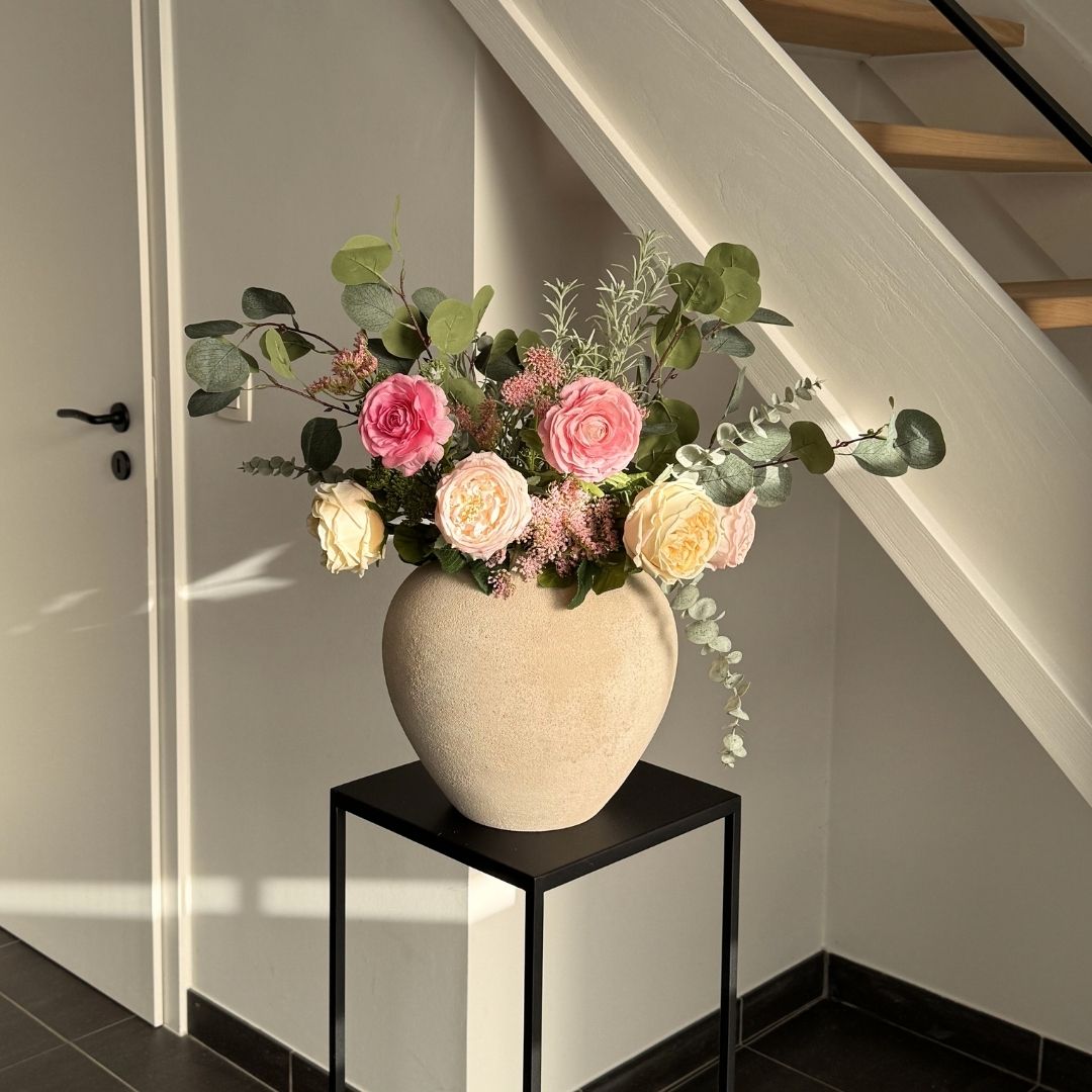 A bouquet of pink and yellow flowers with eucalyptus in a textured beige vase on a black stand.