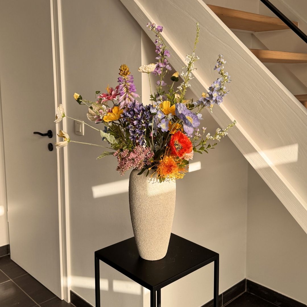 A colorful bouquet of flowers in a textured beige vase sits on a black table in a sunlit corner.