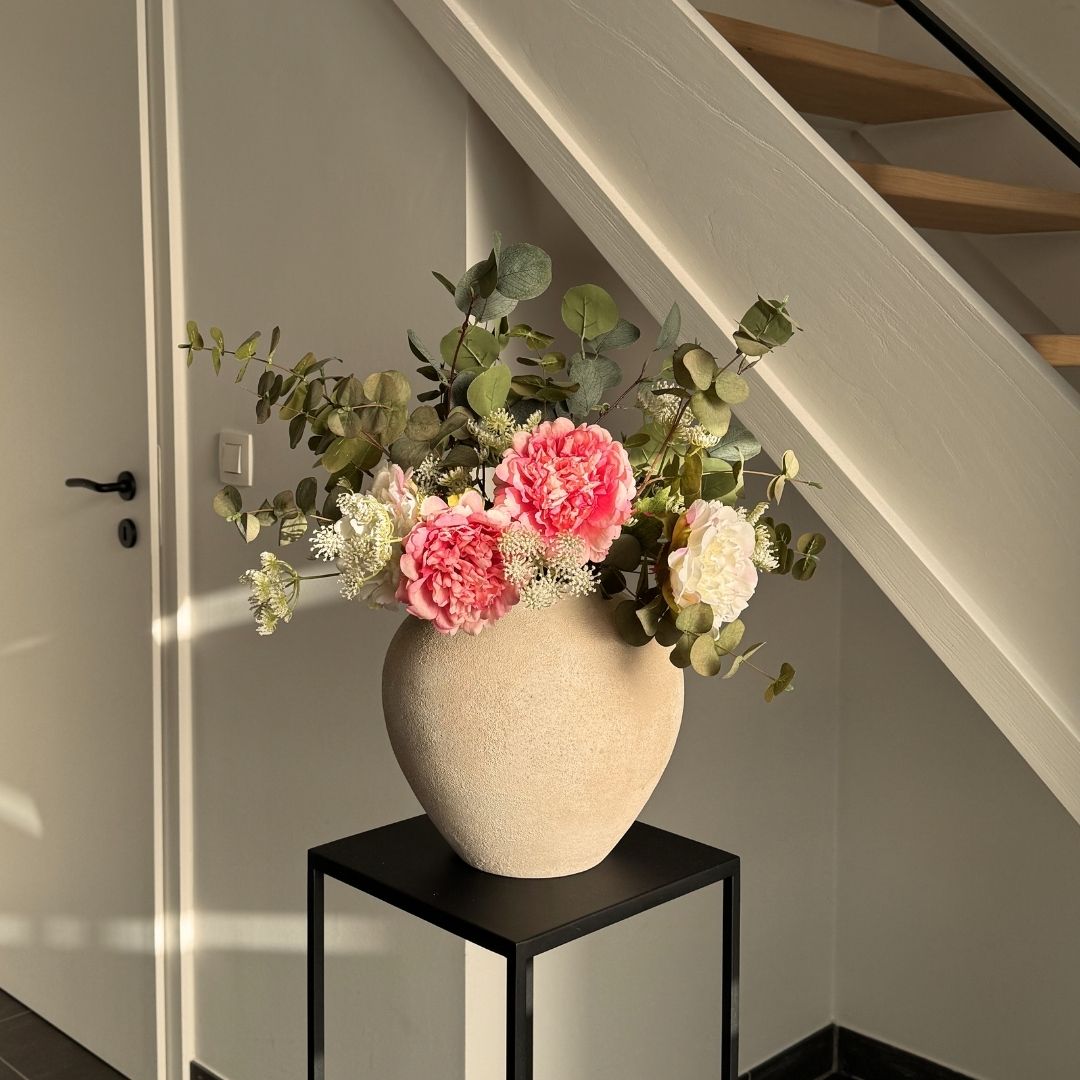 A beige textured vase with pink and white flowers and greenery sits on a black metal stand.