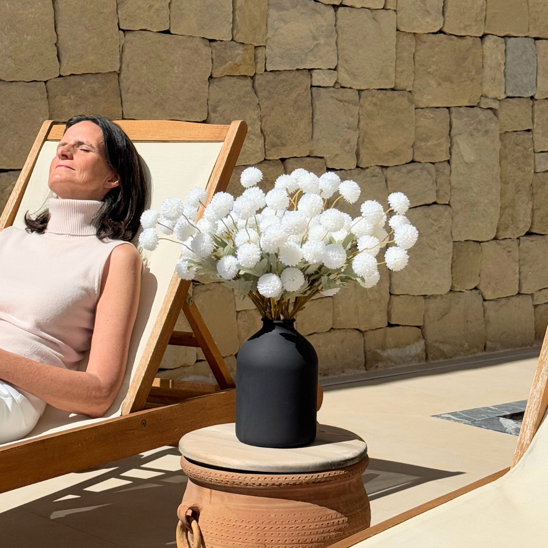 Woman sunbathing on a lounge chair beside a vase of white flowers.
