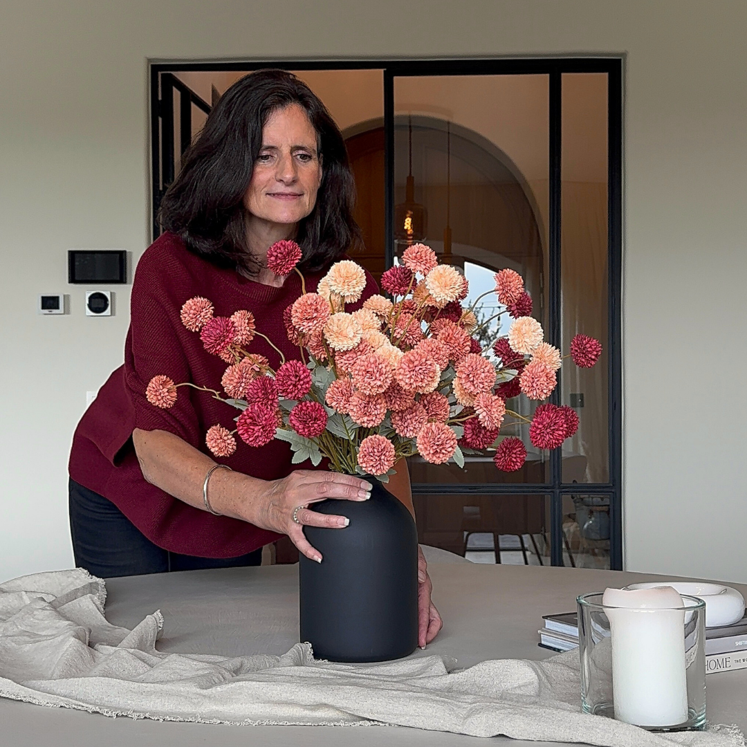 A woman with dark hair arranges a bouquet of pink and peach pom-pom flowers in a black vase.