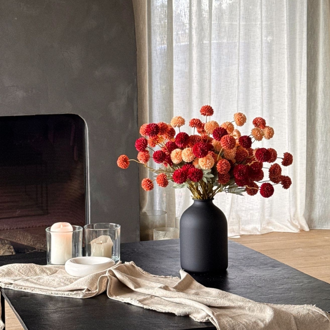 Vase with orange and red flowers on a table, near candles and a fireplace.