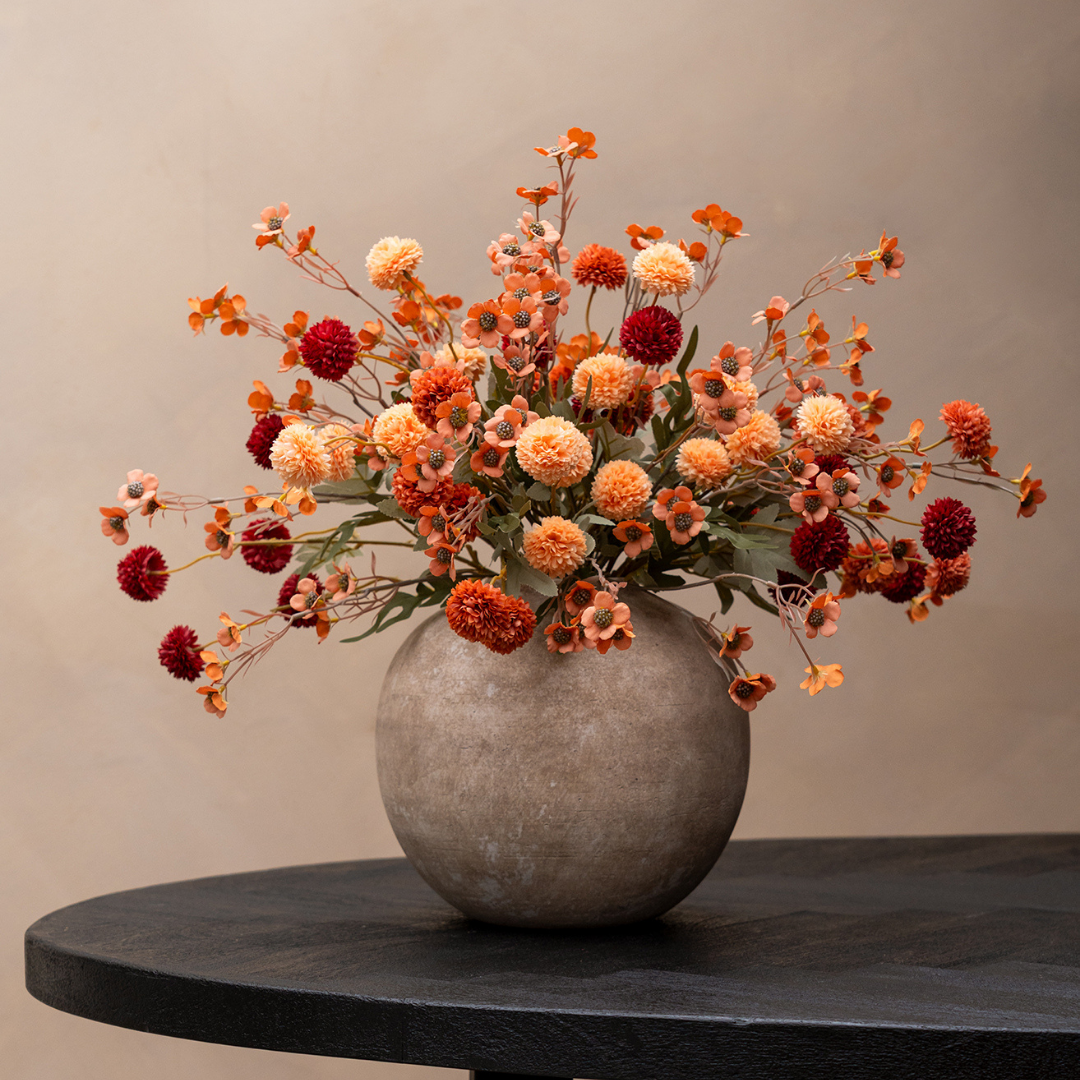 A bouquet of orange and red flowers in a round, grey, textured vase on a dark table.