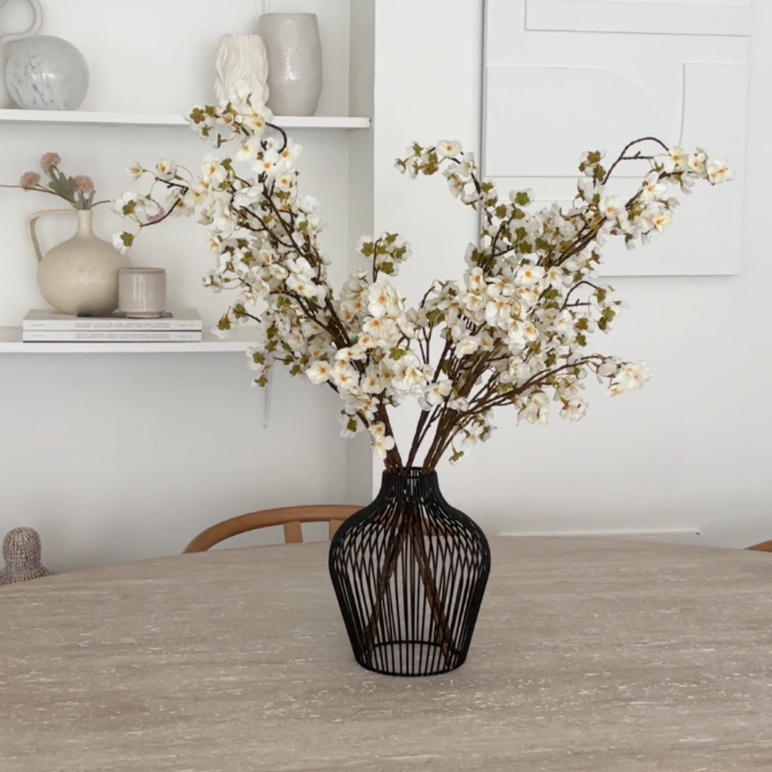 White flowering branches in a black wireframe vase sitting on a light-colored table.