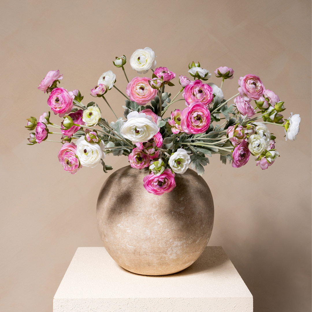 A bouquet of pink and white ranunculus flowers sits in a round, textured vase on a pedestal.