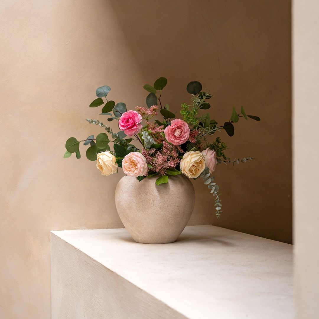 A round vase filled with pink and yellow roses and eucalyptus sits on a white ledge.