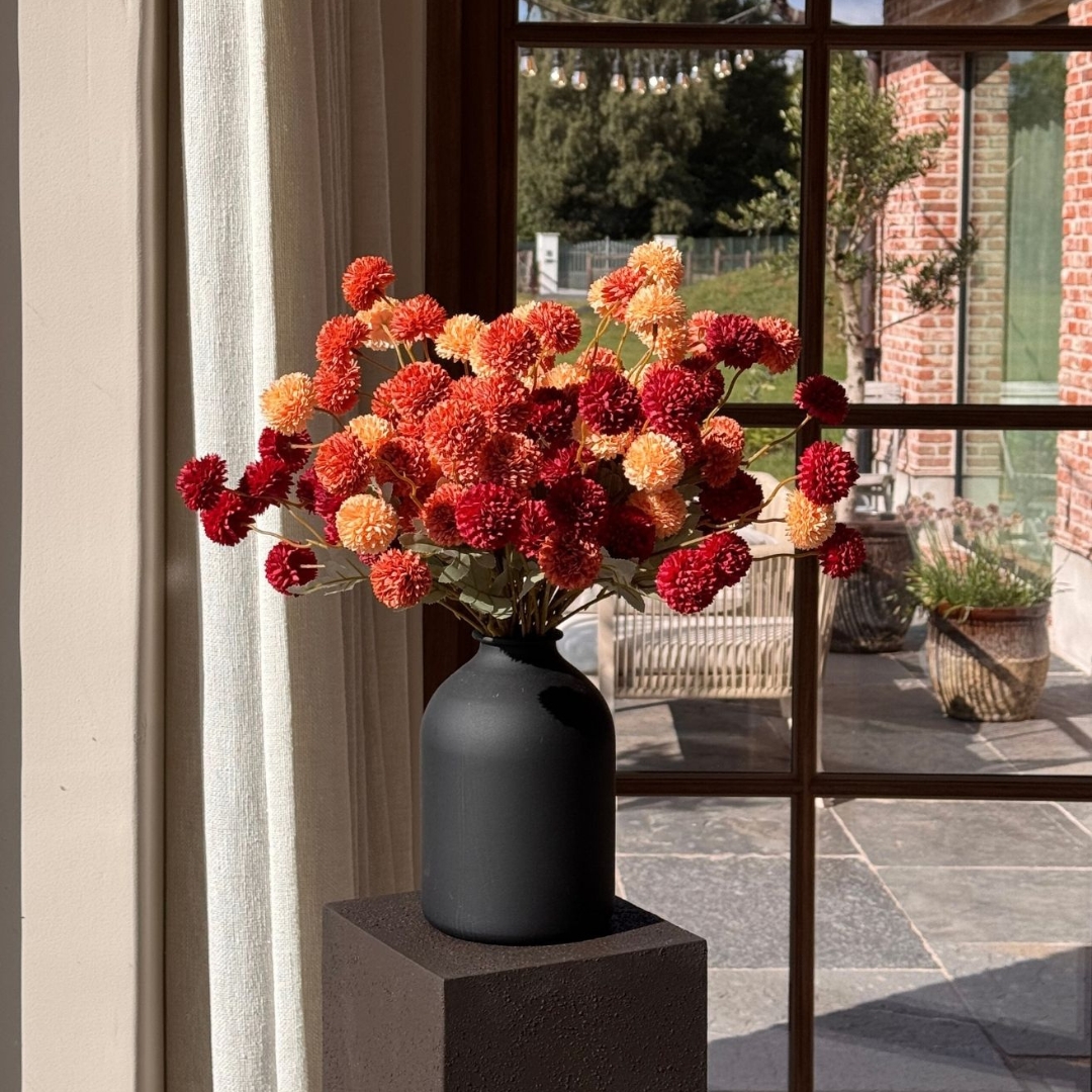 A black vase with orange and red flowers on a pedestal near a window.