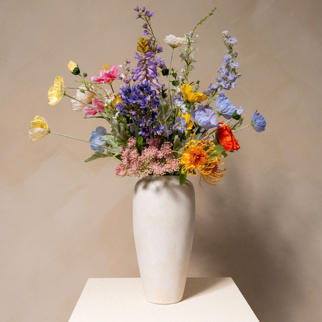 A large, colorful bouquet of various wildflowers in a tall, textured, off-white vase on a pedestal.