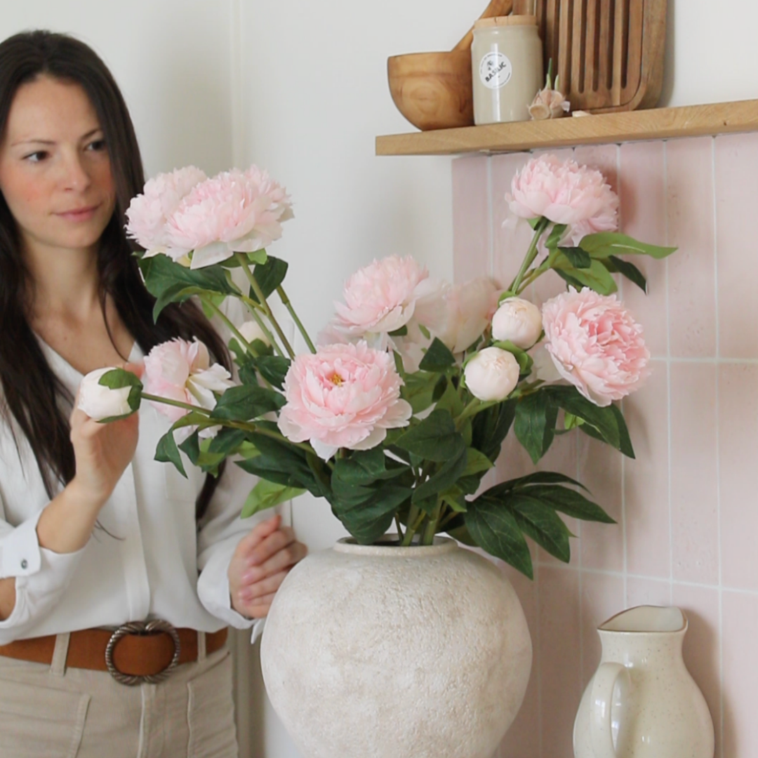 Woman arranging pink peonies in a large vase on a kitchen shelf.