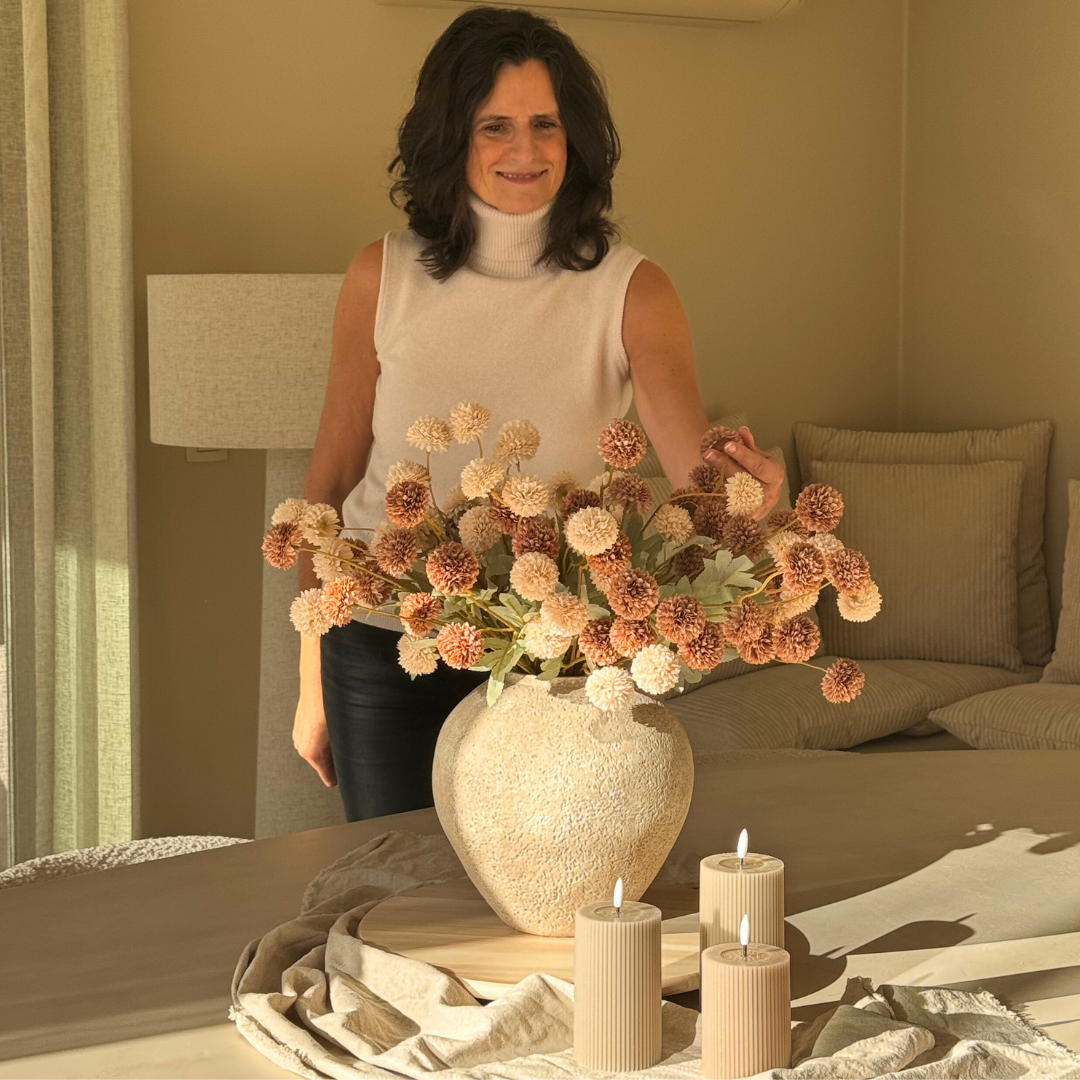 A woman arranges a large bouquet of pom-pom flowers in a textured vase on a table with candles.