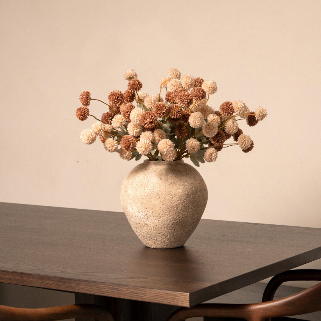 A textured beige vase holding brown and tan pom-pom flowers sits on a dark wooden table.