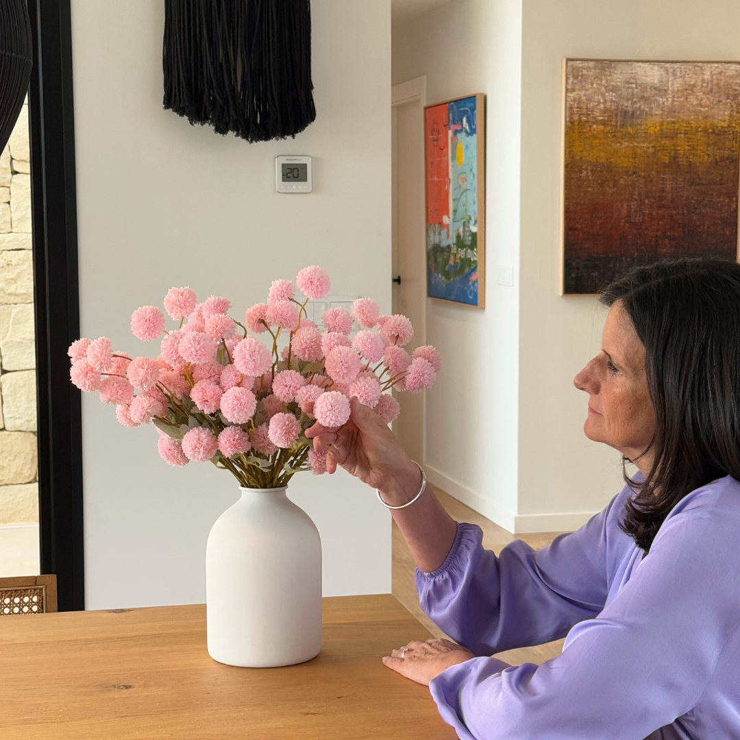 Woman in purple shirt arranging pink flowers in a white vase on a wooden table.