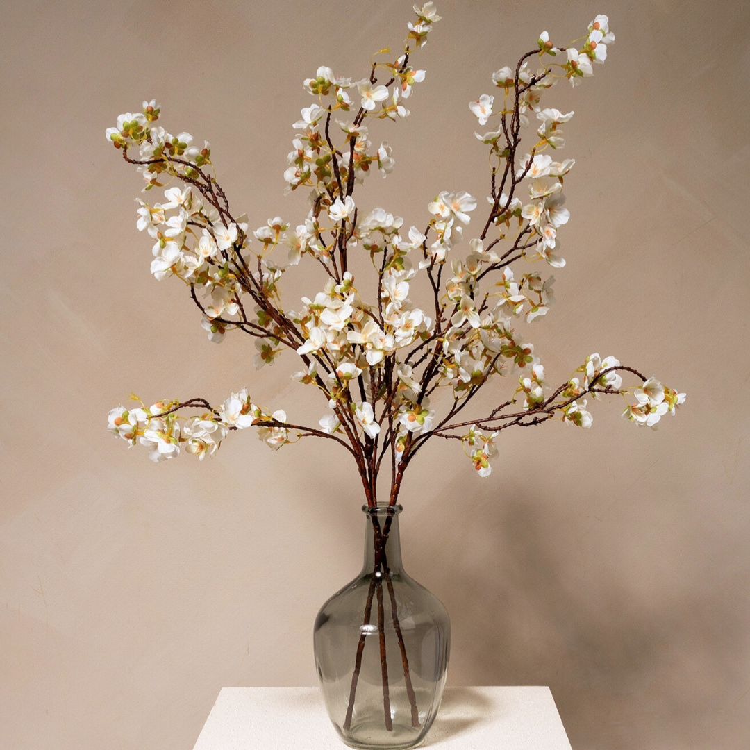 Several branches of white blossoms arranged in a smoked gray glass vase on a white surface.